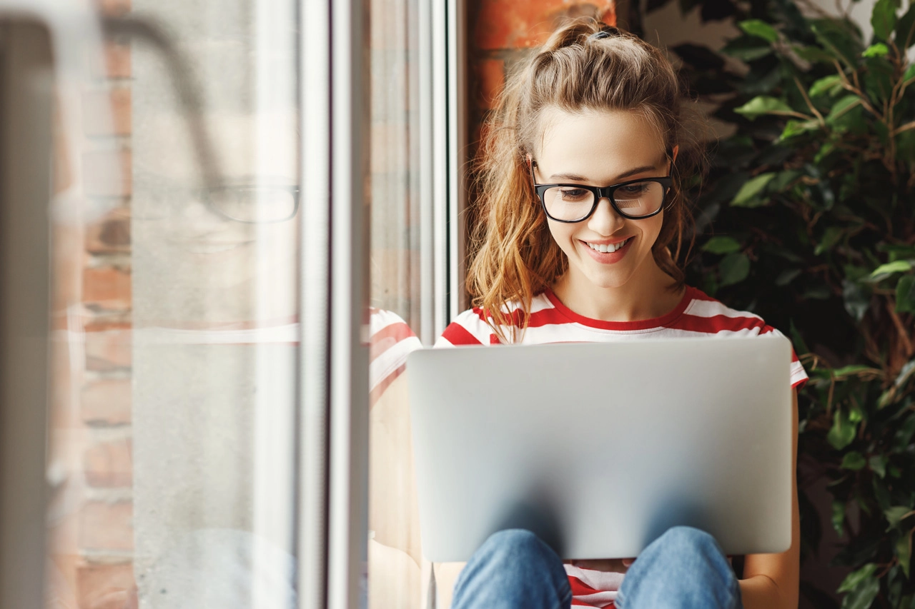 Junge Frau arbeitet lächelnd am Laptop, entspannt am Fenster sitzend.