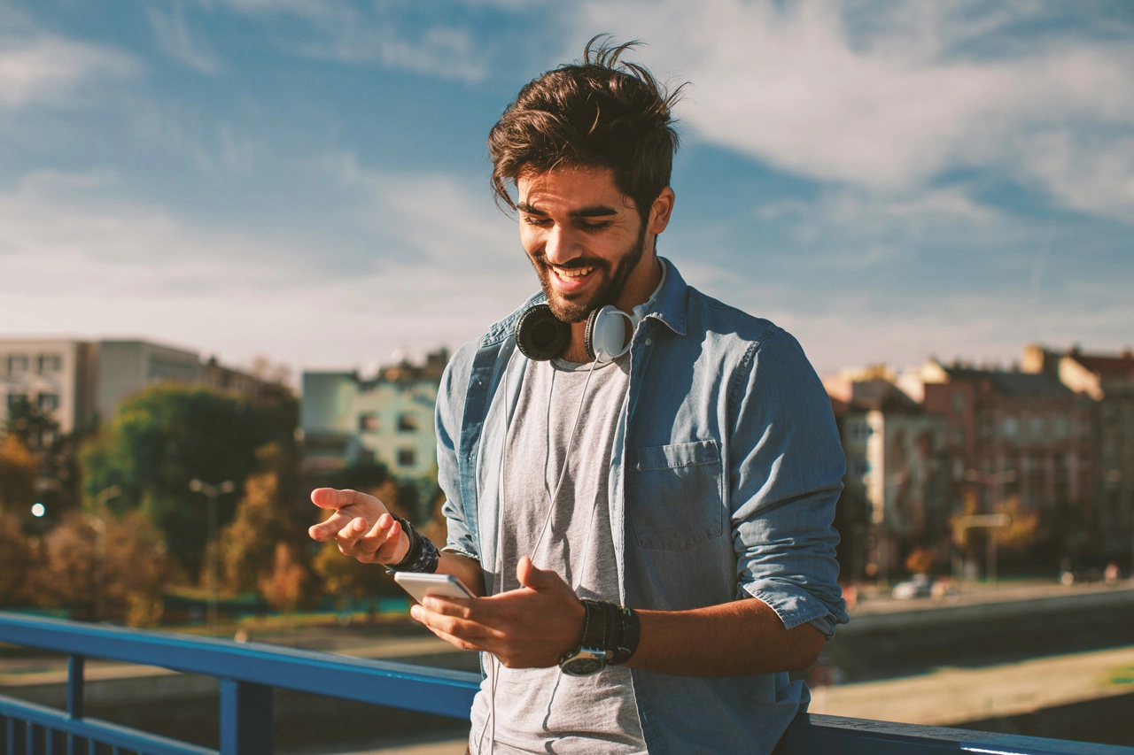 Junger Mann mit Smartphone und Kopfhörern, lächelnd im Freien, urbaner Lifestyle.